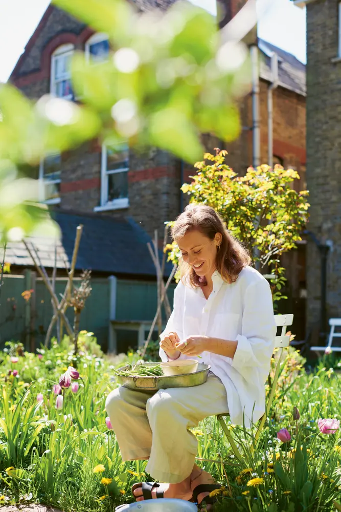 Sophie Gordon sitzt auf einem Stuhl im Garten und sortiert grüne Bohnen mit Haus im Hintergrund.